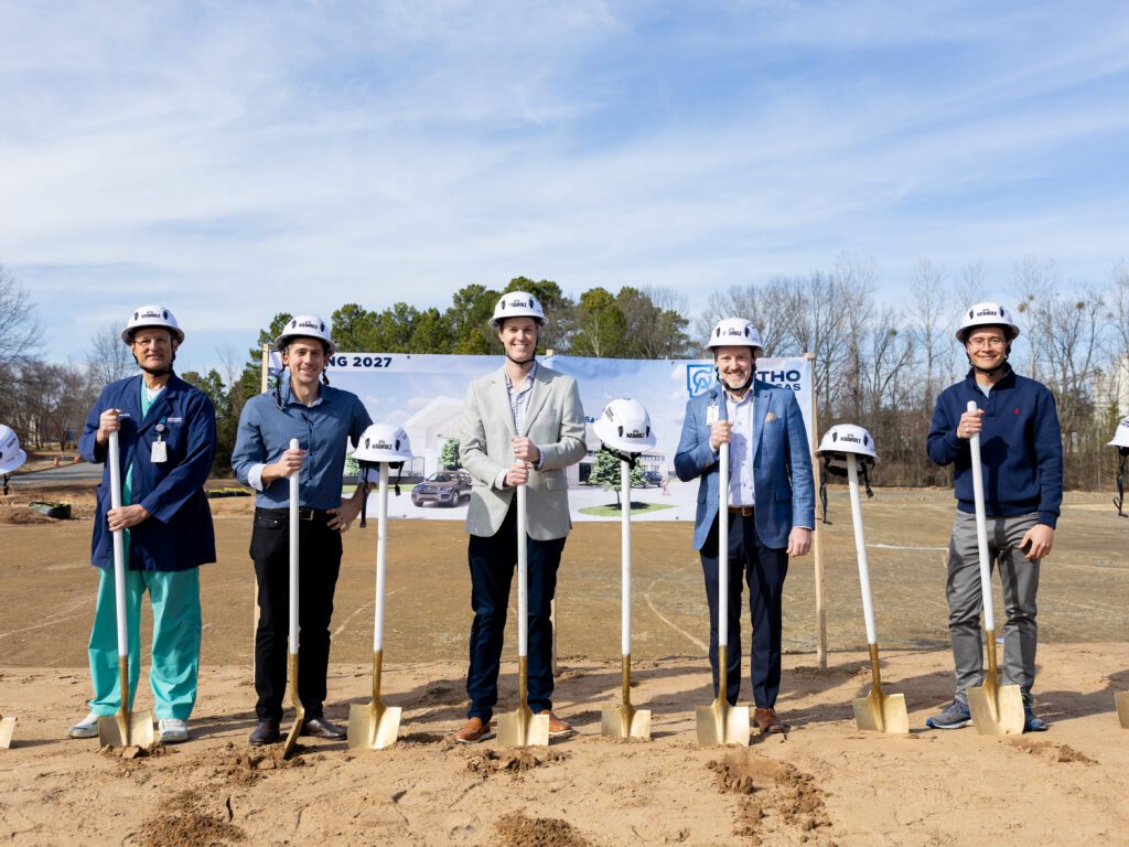 Five men wearing white hard hats stand in a row smiling at the camera, each holding a ceremonial gold shovel in the dirt in front of the OrthoArkansas future site banner. One man on the left is wearing teal surgical scrubs.