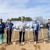 Five men wearing white hard hats stand in a row smiling at the camera, each holding a ceremonial gold shovel in the dirt in front of the OrthoArkansas future site banner. One man on the left is wearing teal surgical scrubs.